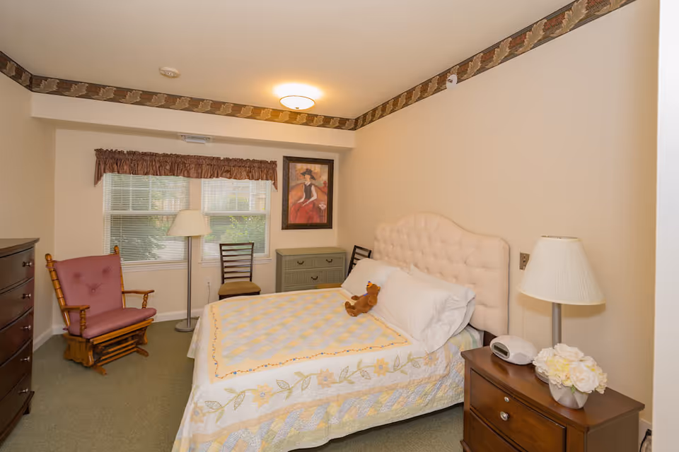 Bright bedroom with a tufted headboard bed covered by a quilt, nightstands with a lamp, a rocking chair, dresser, and a window.
