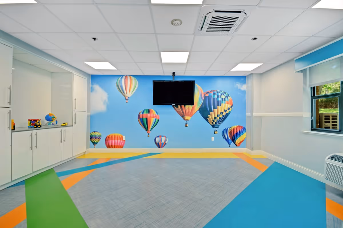 A brightly colored room with a mural of hot air balloons on the far wall. The floor has a geometric pattern with green, orange, blue, and gray sections. There are white cabinets with toys on the left side, a wall-mounted TV in the center, and a window with a view of greenery on the right.