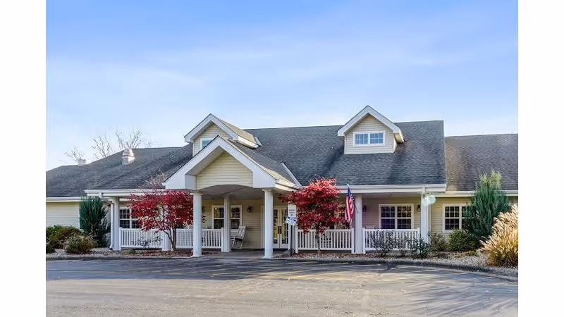 Front entrance of a single-story senior living facility with a covered porch, white railing, and landscaped shrubs and trees.
