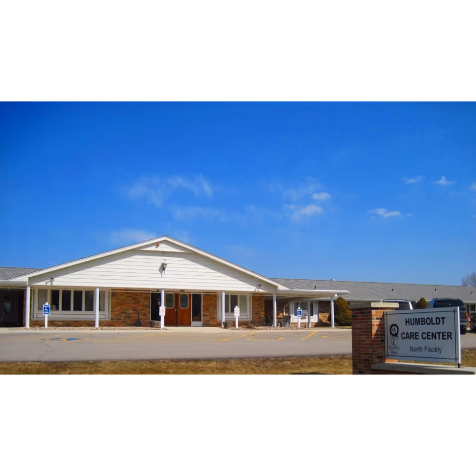 Exterior front view of Humboldt Care Center North, a single-story building with a white gabled roof and brick facade. There is a parking lot with handicap parking spaces in front, and a sign on the right side that reads 'Humboldt Care Center North Facility'. The sky is clear and blue.