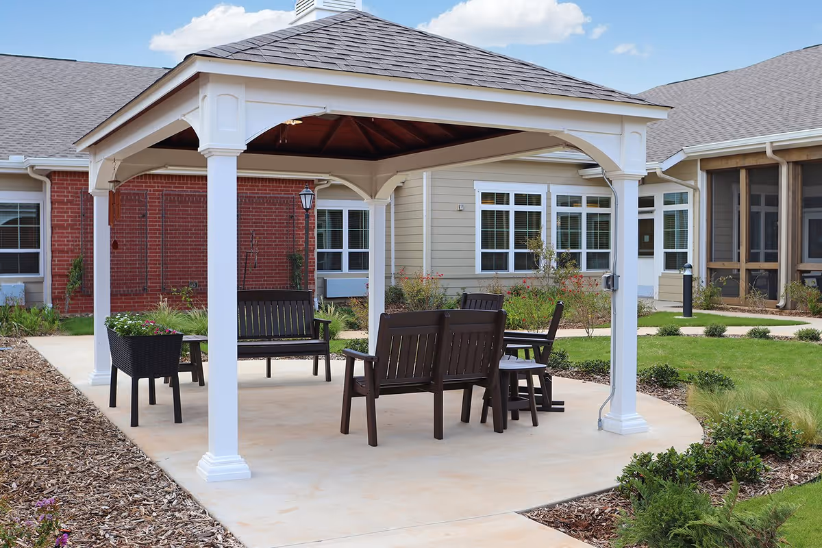 Outdoor seating area with a white gazebo covering several dark wooden benches and chairs, surrounded by landscaped garden beds and grass, with a building featuring windows and a screened porch in the background under a partly cloudy sky.