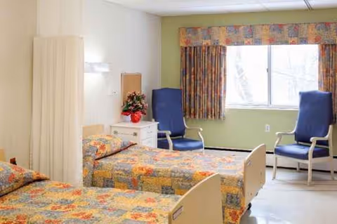 Two beds with patterned quilts, a nightstand, and blue chairs by a window.