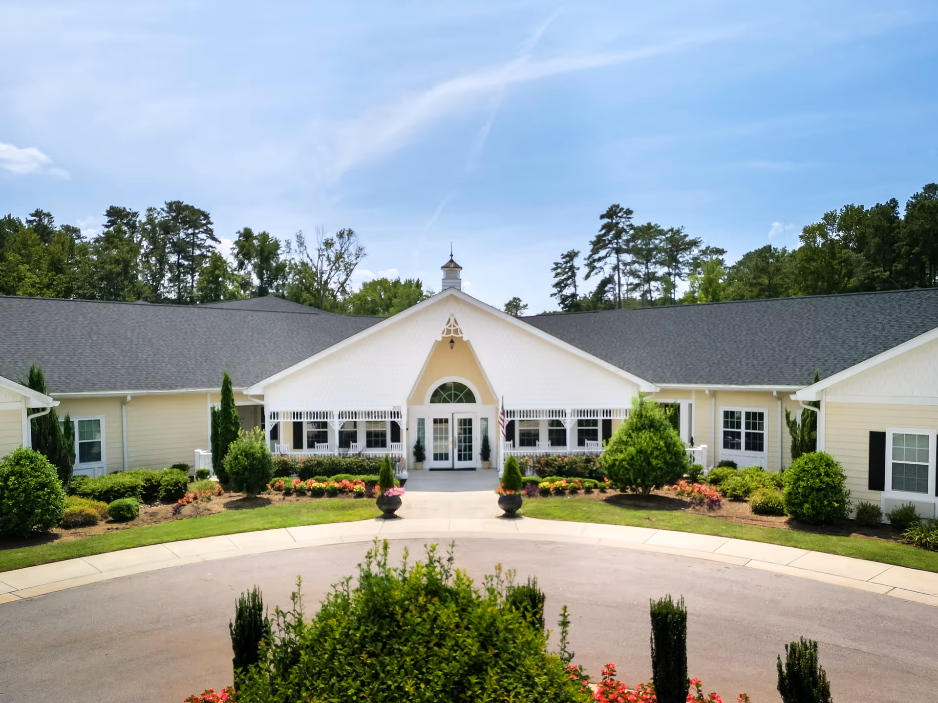 Front exterior view of a single-story senior living facility building named Cadence Garner, featuring a central entrance with white double doors, surrounded by well-maintained landscaping including bushes, flowers, and trees under a clear blue sky.