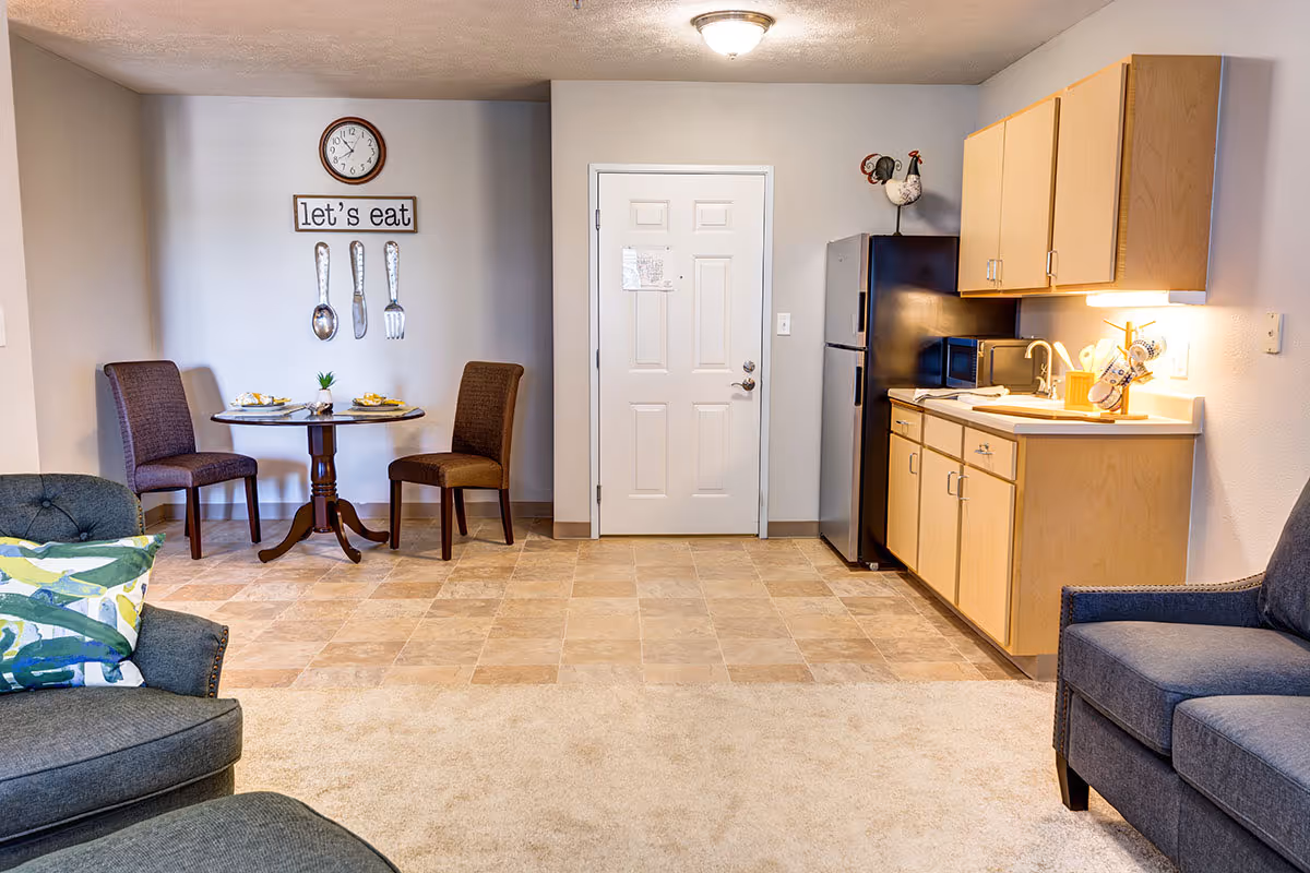 A small dining area and kitchenette in a senior living facility. The dining area has a round wooden table with two brown upholstered chairs and place settings. Above the table is a wall clock and a decorative sign that says 'let's eat' with large spoon, knife, and fork wall hangings. The kitchenette features light wood cabinets, a black refrigerator, a microwave, a sink, and various kitchen utensils. Two gray upholstered chairs with patterned pillows are partially visible in the foreground.