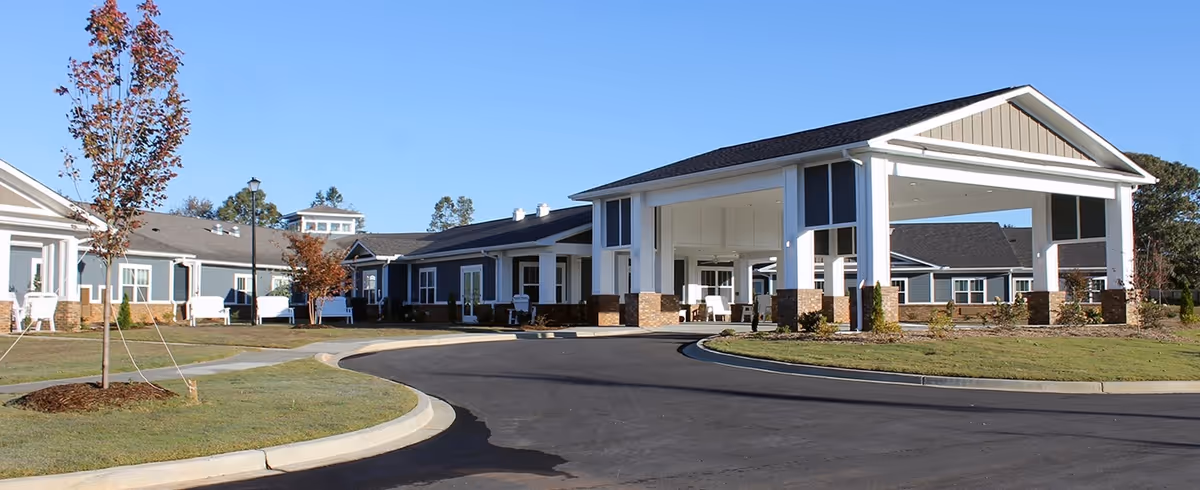 Exterior view of Lakewood Assisted Living facility on Clark Road in Boiling Springs, SC, showing a large covered entrance with white columns and brick bases, surrounded by a curved driveway, landscaped grass, and small trees under a clear blue sky.