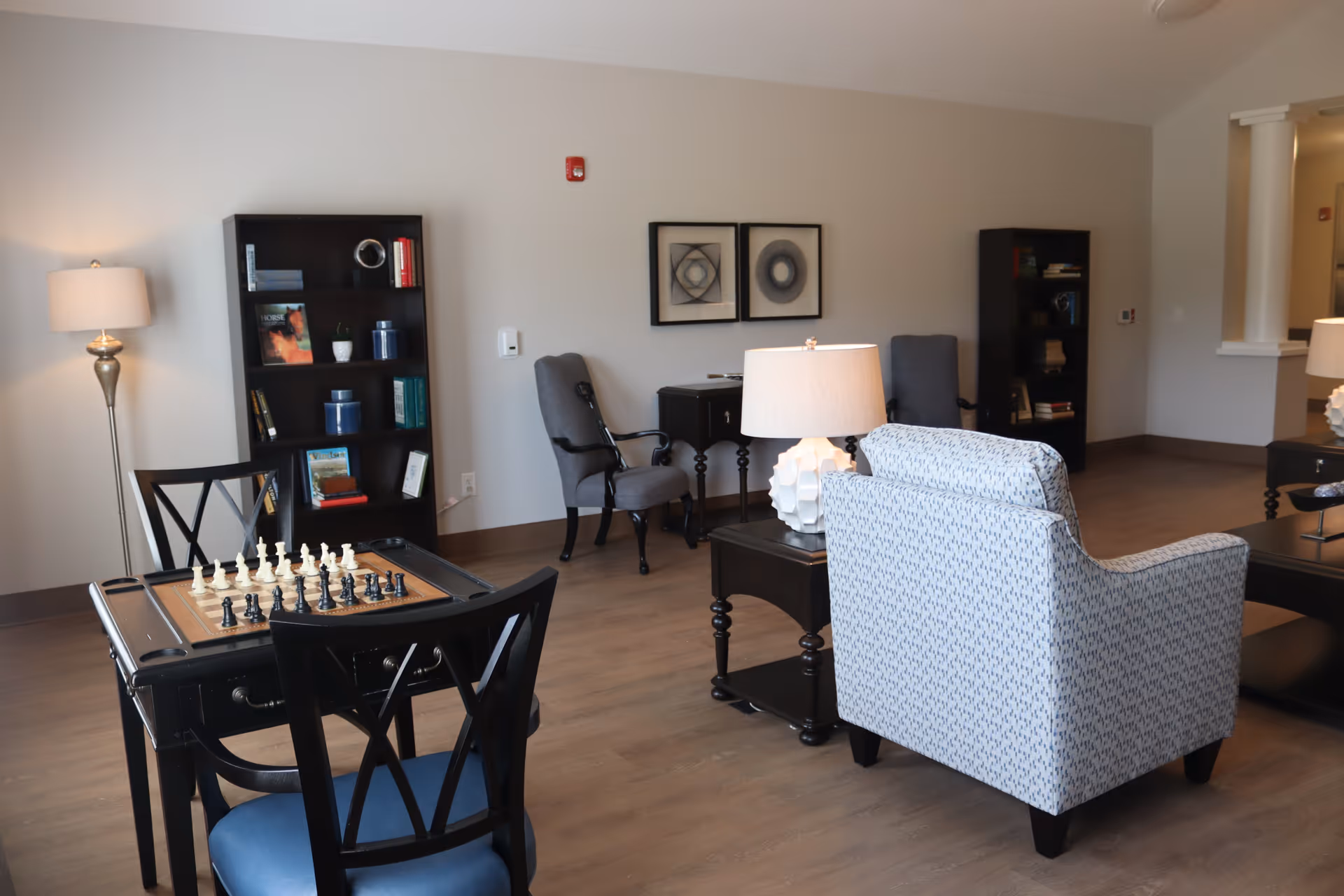 A bright common living room with armchairs, a chess table, bookshelves and lamps.