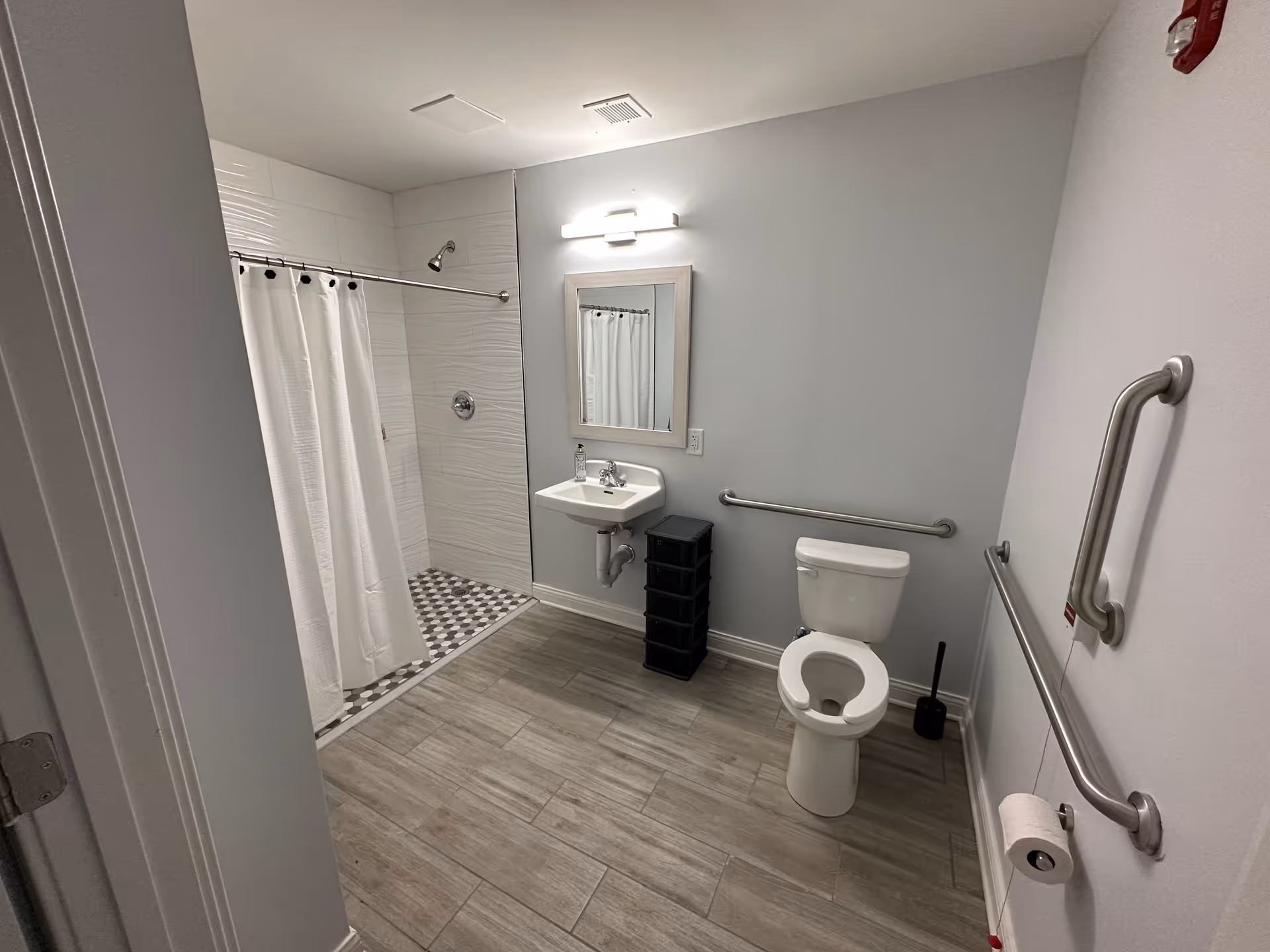 A clean, accessible bathroom with a walk-in shower featuring a white curtain, a wall-mounted sink with a mirror above it, a toilet with grab bars on the adjacent walls, and a toilet paper holder. The floor has light wood-like tiles and the walls are painted light gray.