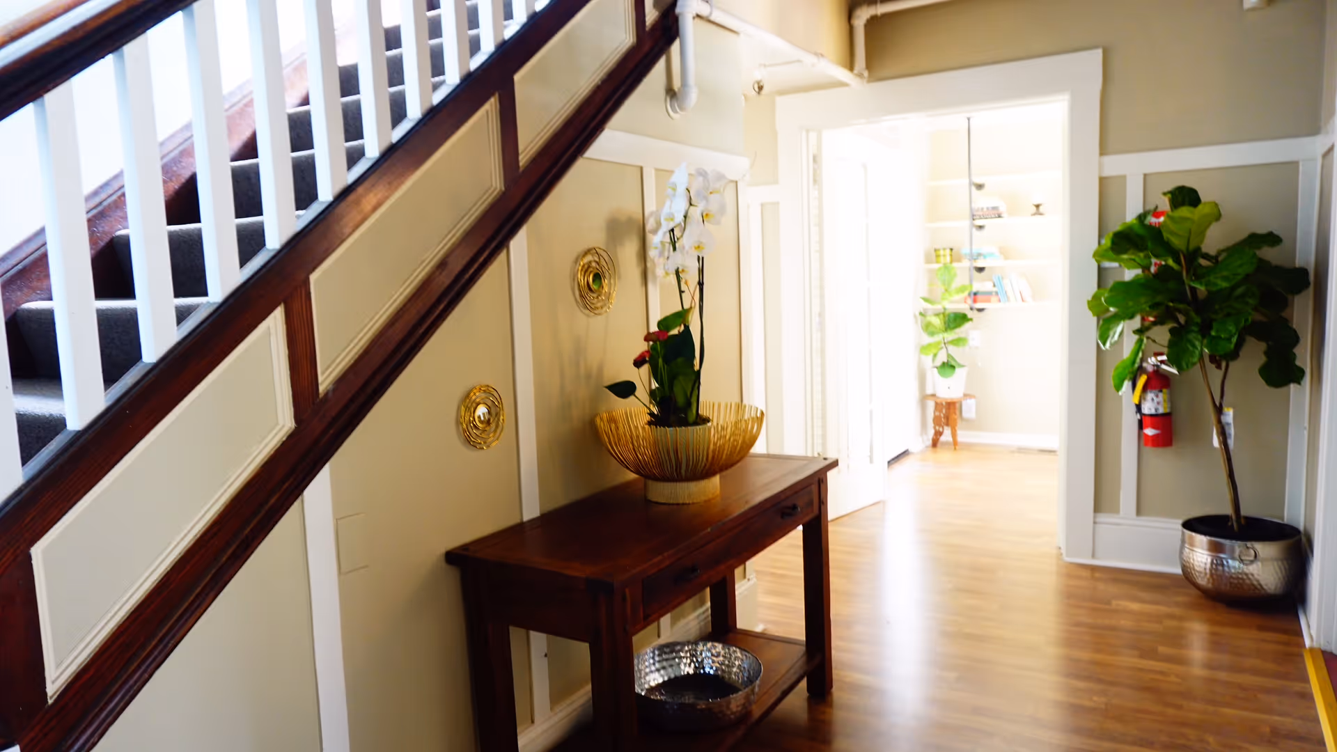 Interior hallway with wooden flooring and a staircase with white railings on the left. A wooden console table with a decorative bowl and a white orchid plant is placed against the wall under the staircase. The hallway leads to a bright room with shelves and plants. A large potted plant and a fire extinguisher are visible on the right side of the hallway.