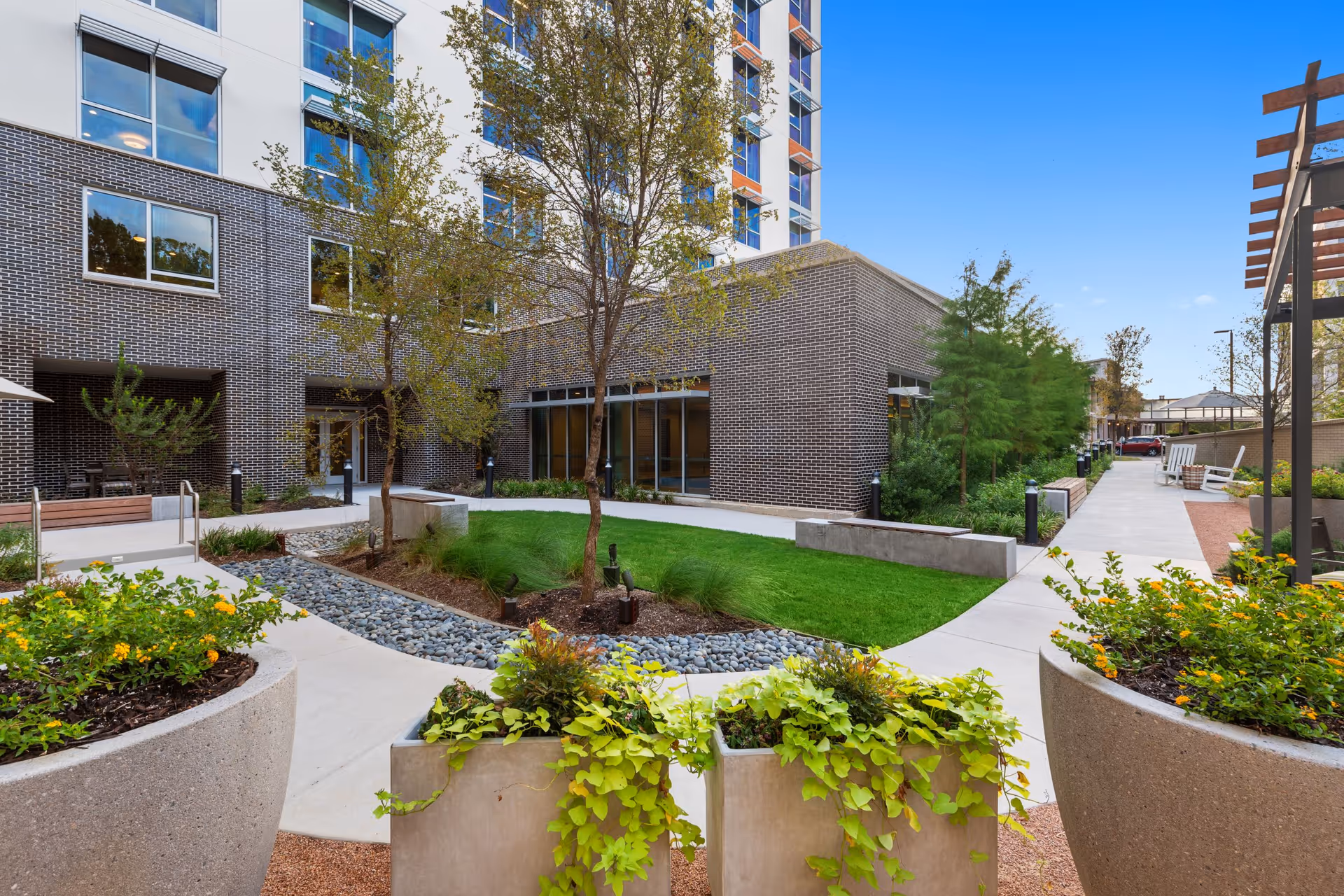 Outdoor courtyard area of a senior living facility with concrete walkways, green grass, trees, and large planters filled with various plants and flowers. The building exterior features brick and multiple windows, with benches and seating areas along the pathways under a clear blue sky.