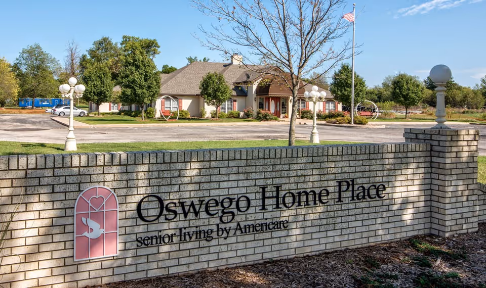 Exterior view of Oswego Home Place Senior Living facility with a brick sign in the foreground displaying the facility name and logo, a parking lot, trees, and a building with a pitched roof in the background under a clear blue sky.