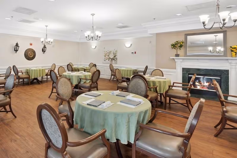 A dining room with several round tables covered with green tablecloths, each set with napkins and menus. The room has wooden floors, upholstered chairs, a fireplace with a mirror above it, and soft lighting from chandeliers and wall sconces.