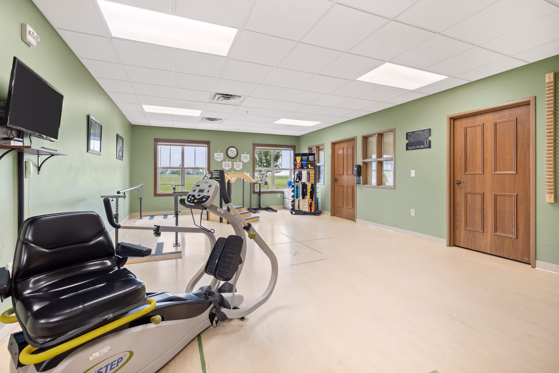 A spacious exercise room with light green walls and light wood flooring. The room contains exercise equipment including a recumbent bike and parallel bars. There are two large windows letting in natural light, a wall-mounted TV, and various exercise accessories organized on a rack near the windows. The ceiling has recessed lighting, and there are two wooden doors on the right wall.