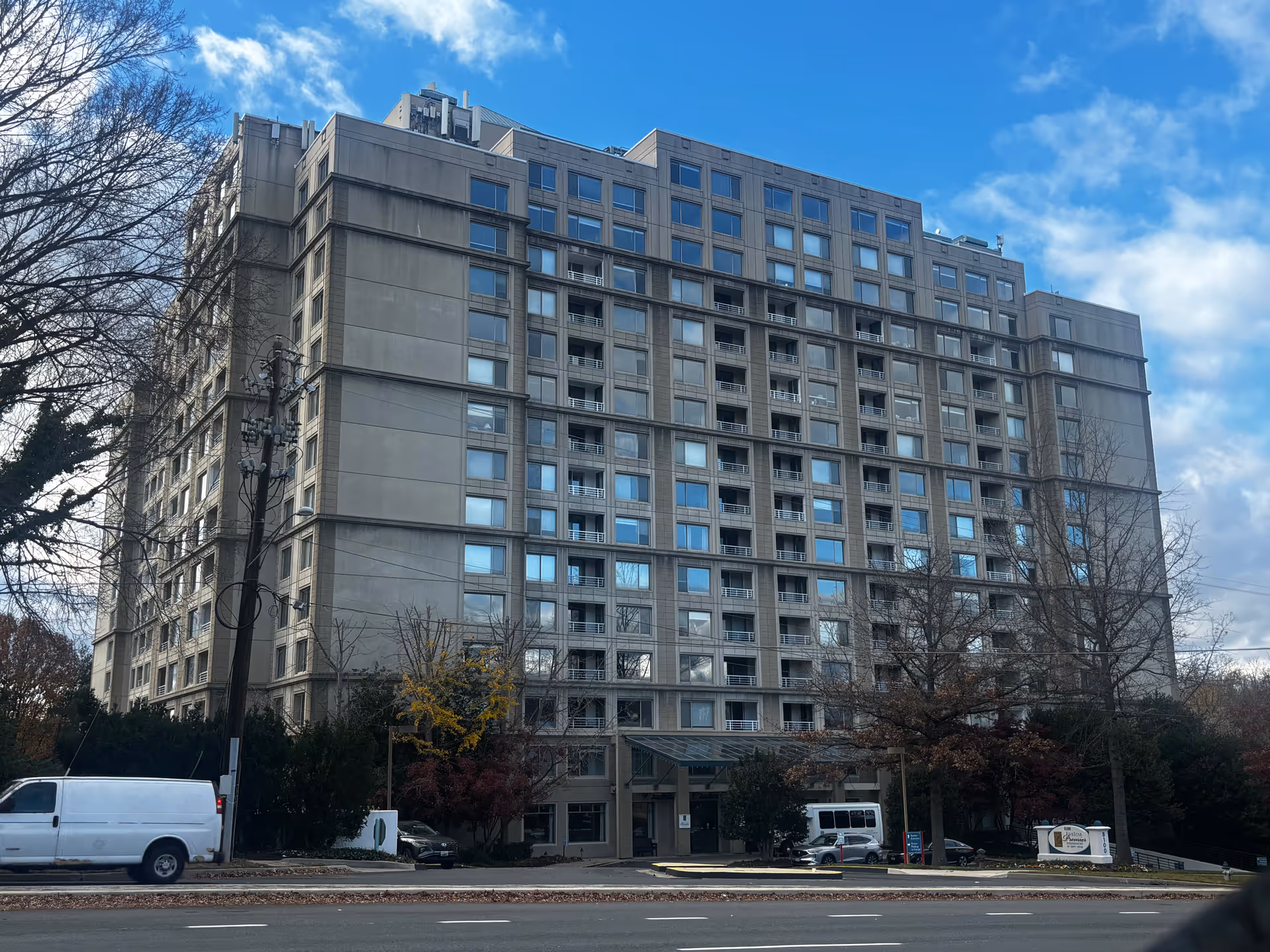 Exterior view of a multi-story senior living facility building named Five Star Premier Residences of Chevy Chase, with multiple windows and a covered entrance. There are trees with autumn foliage and several vehicles parked in front of the building under a partly cloudy blue sky.