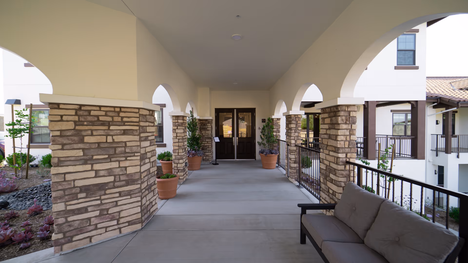 Covered outdoor walkway with stone pillars and arched openings leading to double doors. Potted plants line the walkway and a cushioned bench is on the right side. The building exterior is visible with windows and railings.