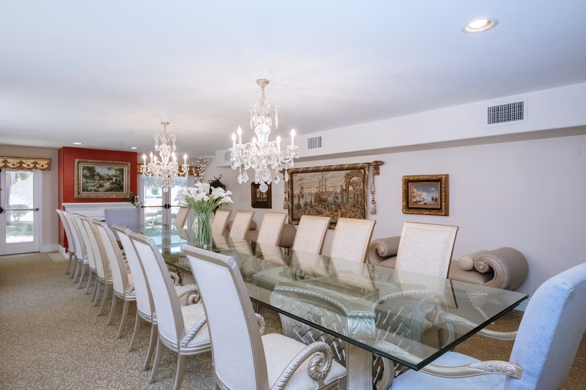 A large, elegant dining room with a long glass table surrounded by twelve upholstered chairs. Two ornate crystal chandeliers hang above the table. The room features framed artwork on the walls, a red accent wall with a painting above a white fireplace, and large windows with decorative valances letting in natural light.