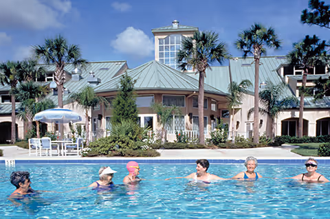 Six older adults in a swimming pool in front of a palm-tree-lined senior living building under a blue sky.