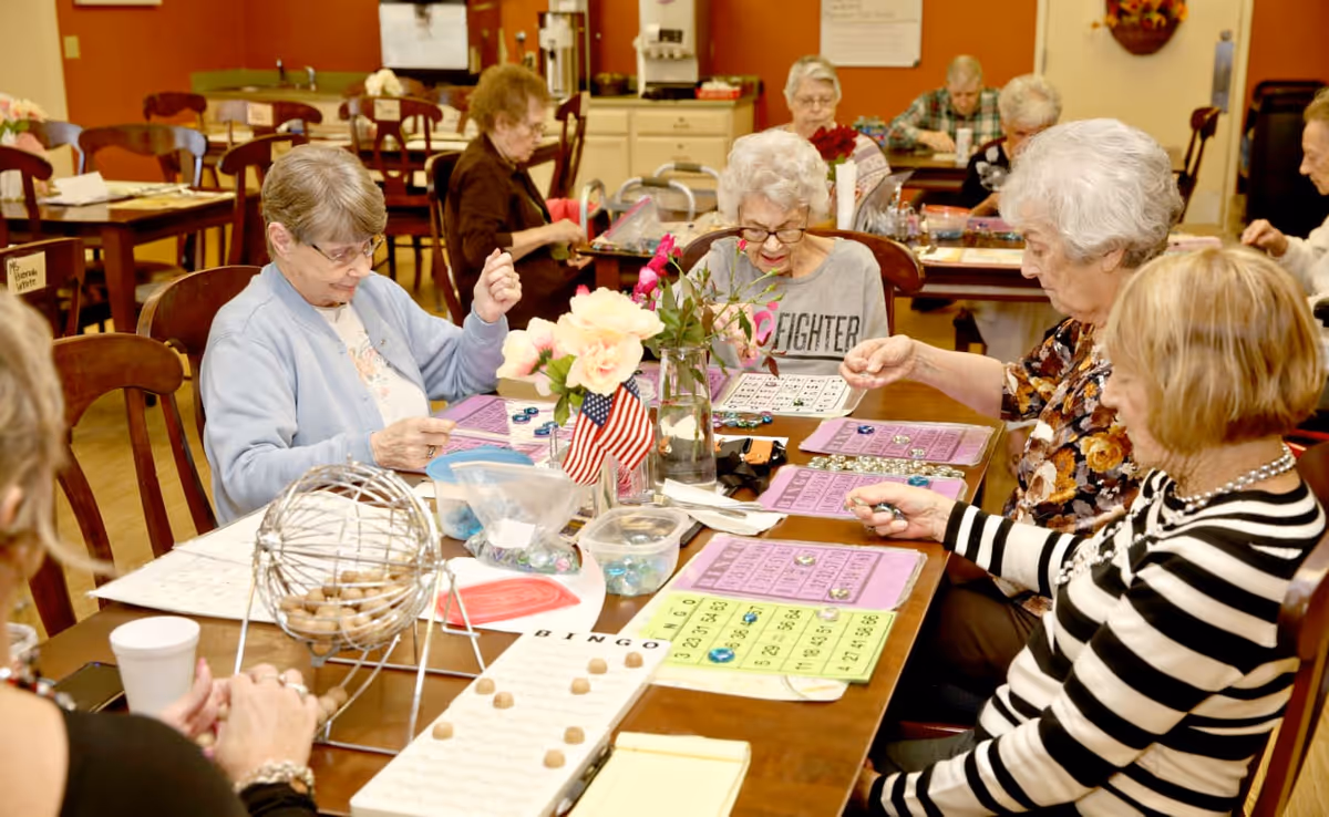 A group of elderly women sitting around a table playing bingo in a communal room. The table is covered with bingo cards, markers, and a bingo cage with balls. The room has warm lighting and a cozy atmosphere with other tables and chairs in the background.