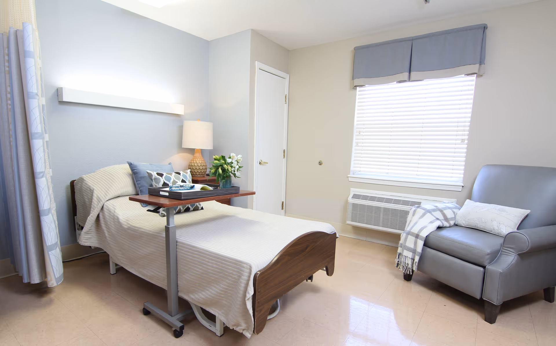 A clean and bright nursing facility room with a single bed covered in a beige blanket, a small overbed table with a book and a tray, a bedside lamp, and a vase of flowers. There is a comfortable gray armchair with a plaid throw and a pillow next to a window with blinds and a blue valance. The room has light-colored walls and flooring.