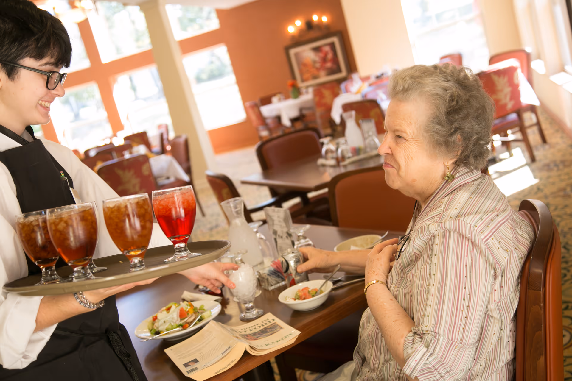 A young waiter serving drinks to an elderly woman seated at a dining table in a well-lit dining room. The table has plates of food, a newspaper, and a glass of ice water. The room has large windows and several tables with chairs.