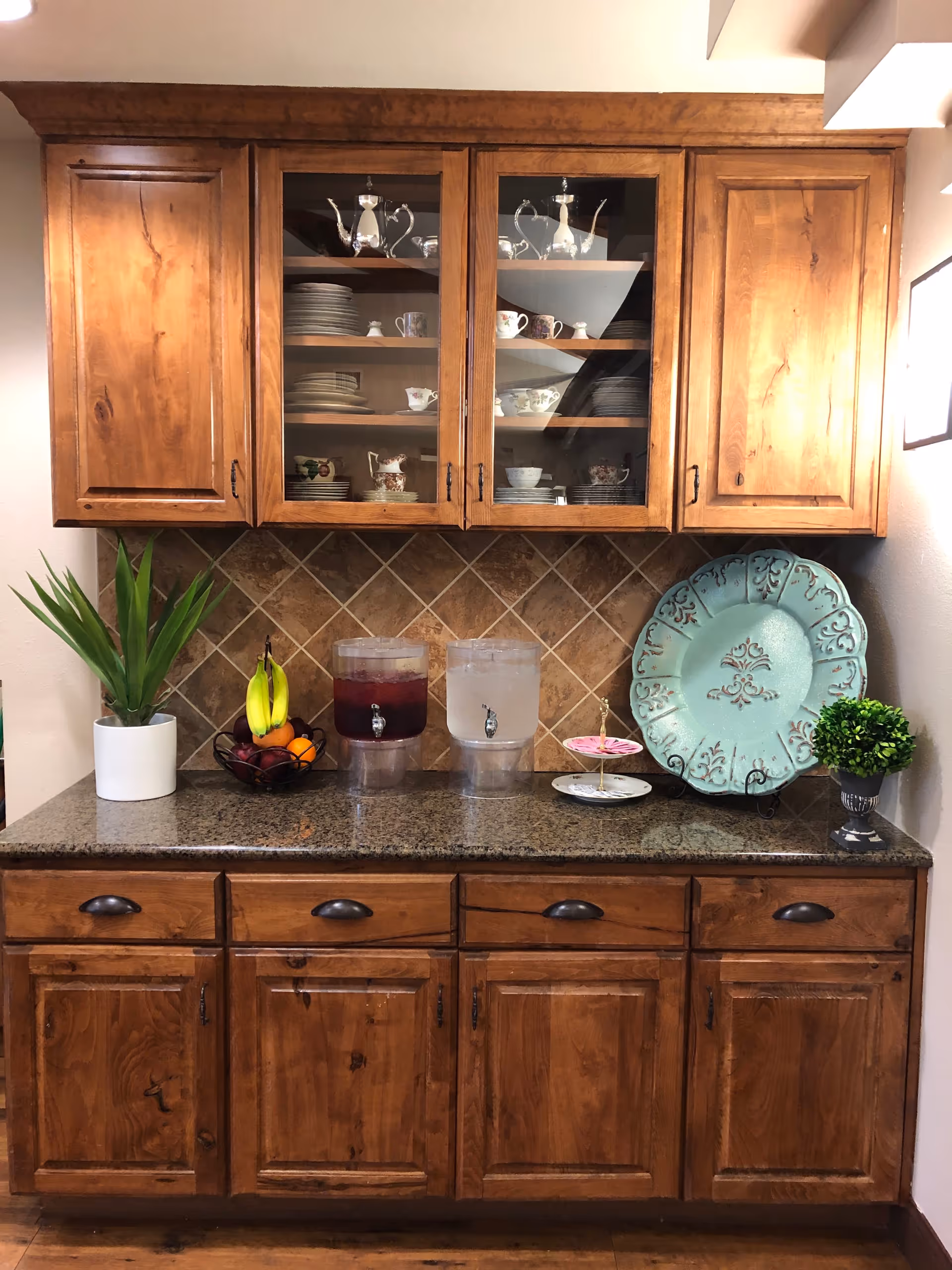 Wooden kitchen cabinet with glass doors displaying china and silver tea sets. Below the cabinet is a granite countertop with two beverage dispensers, a bowl of fruit, a potted plant, a decorative blue plate on a stand, and a small green plant in a pot. The backsplash is made of brown tiles arranged in a diamond pattern.