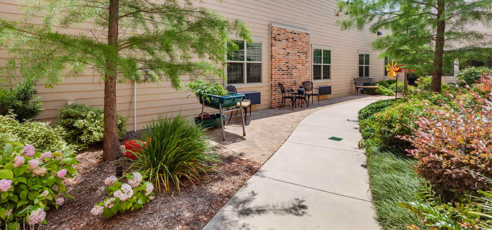 A landscaped outdoor walkway alongside a building with beige siding and brick accents. The path is bordered by green shrubs, flowering plants, and trees. There are several black metal chairs and small tables placed on a paved area next to the building.