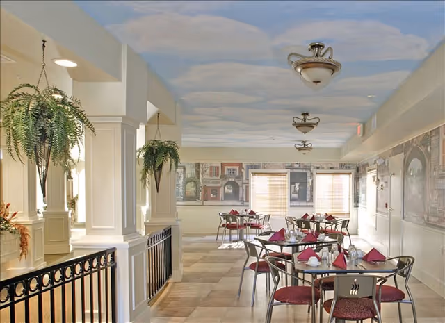 Dining room with round tables set with napkins and chairs beneath a painted sky ceiling and hanging plants.