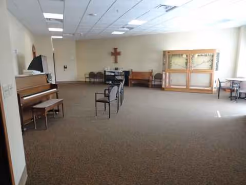 Spacious senior facility common room with chairs, piano, and a cross on the wall.
