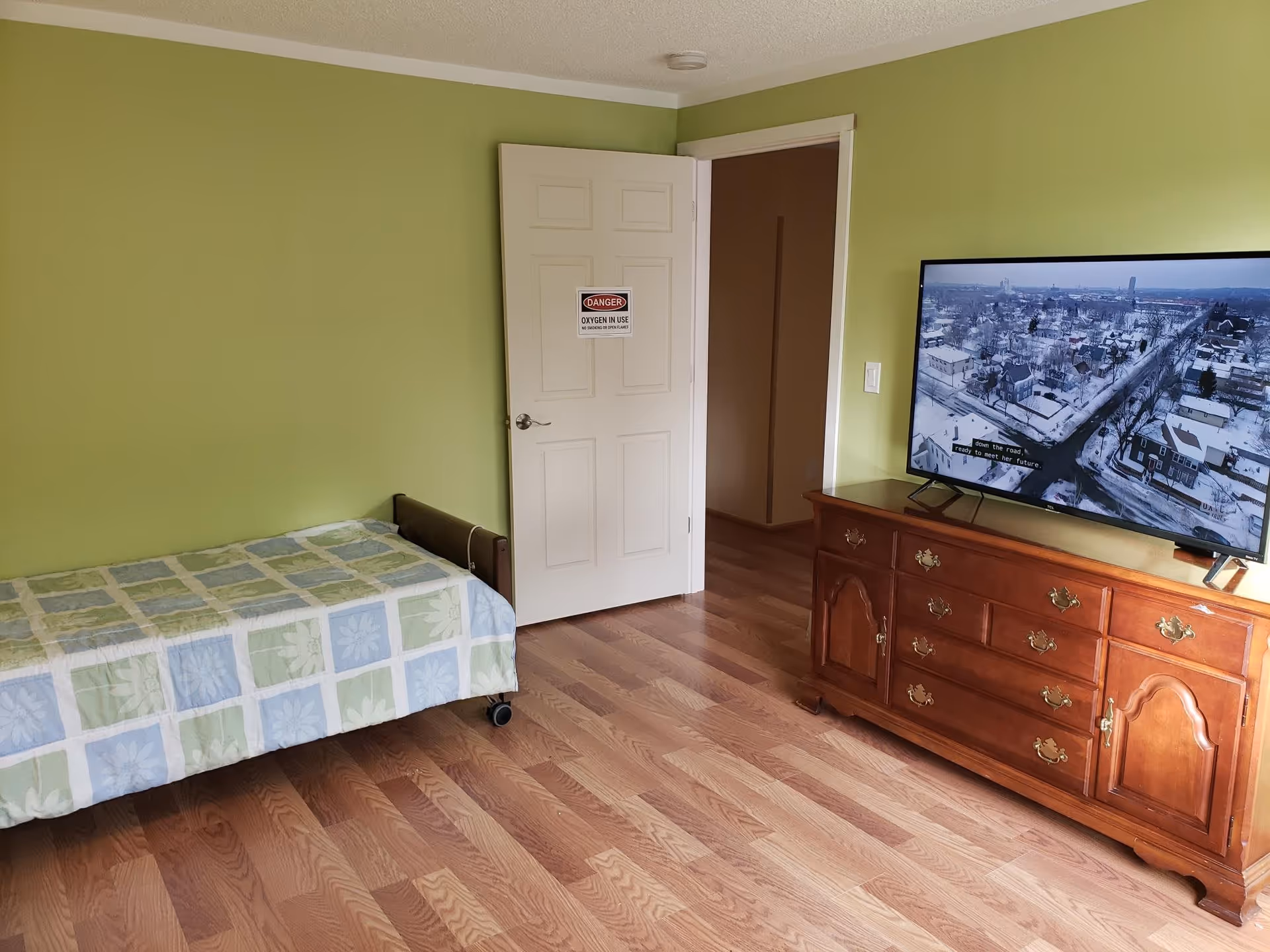 A bedroom with light green walls, a single bed with a patterned bedspread, a wooden dresser with brass handles, and a flat-screen TV displaying an aerial view of a snowy town. The door to the room is open and has a sign that reads 'DANGER OXYGEN IN USE NO SMOKING OR OPEN FLAMES'.
