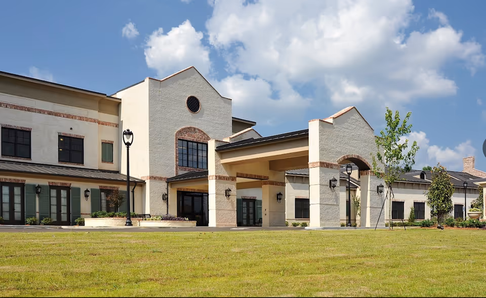 Exterior view of a large, light-colored senior living facility building with brick accents, multiple windows, and a covered entrance. The building is surrounded by a well-maintained lawn and a few young trees under a partly cloudy blue sky.