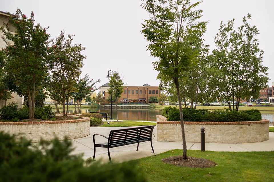 Park-like pond area with benches, trees, and a walkway in front of a brick building across the water.
