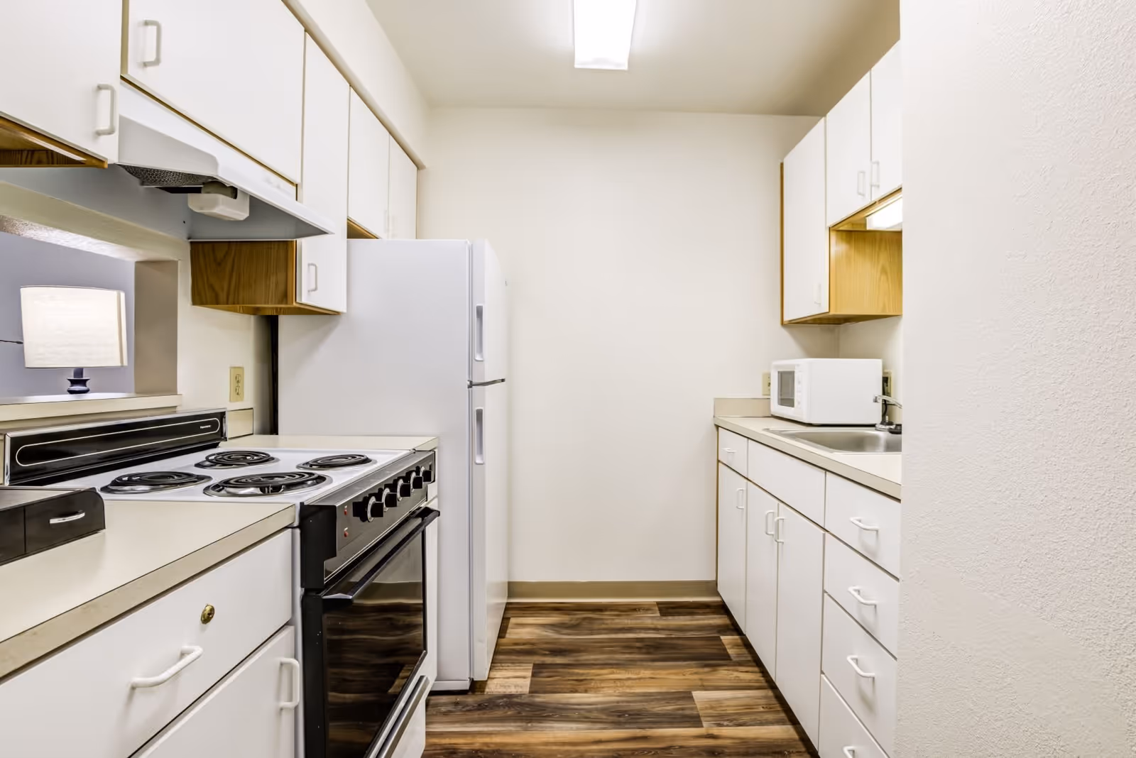 A compact kitchen with white cabinets, a white refrigerator, an electric stove with coil burners, a microwave on the countertop, a sink, and wood-patterned flooring. A lamp is visible through a pass-through window above the stove.