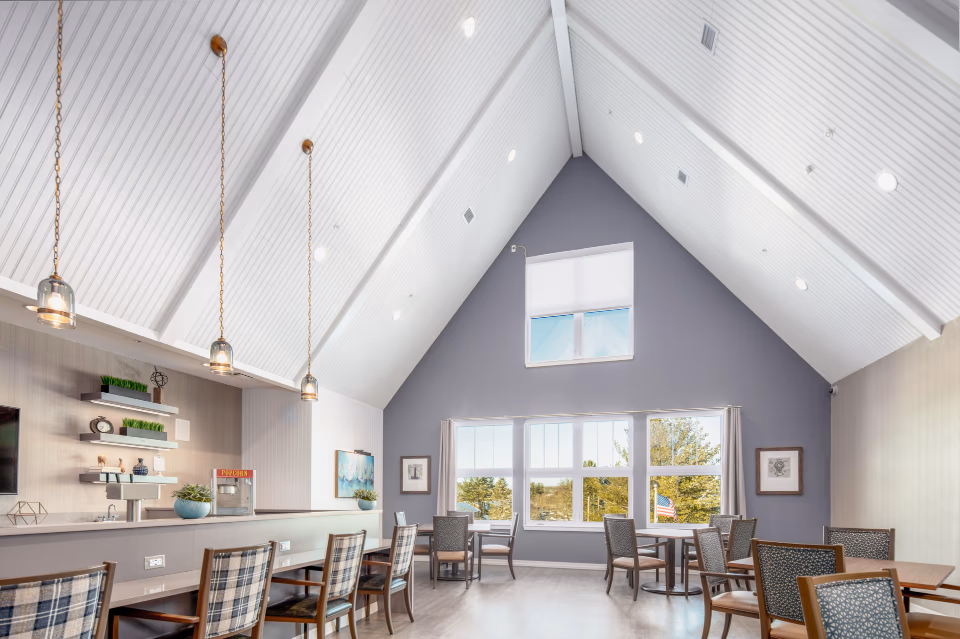 A bright and spacious dining area with high vaulted white ceilings and large windows letting in natural light. The room features several tables and chairs arranged for dining, a counter with bar stools, pendant lights hanging from the ceiling, and decorative shelves with plants and ornaments. The walls are painted in soft gray tones, and there is a popcorn machine on the counter.
