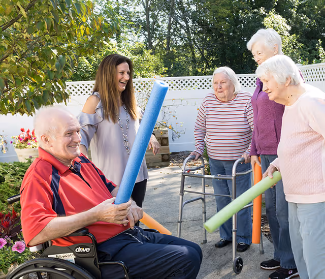 A group of elderly people and a caregiver are outdoors in a garden area. One elderly man in a wheelchair is holding a blue pool noodle, while three elderly women, one using a walker, and the caregiver stand around him smiling and holding colorful pool noodles. They appear to be enjoying a light exercise or recreational activity together.