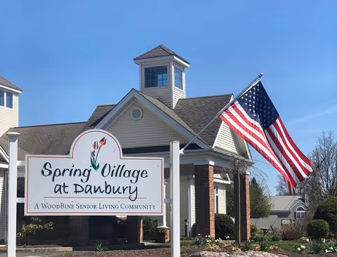 Exterior view of Spring Village at Danbury senior living community building with a large American flag mounted on a pole. A white sign in front reads 'Spring Village at Danbury, A Woodbine Senior Living Community'. The building has beige siding, brick columns, and a small cupola on the roof under a clear blue sky.