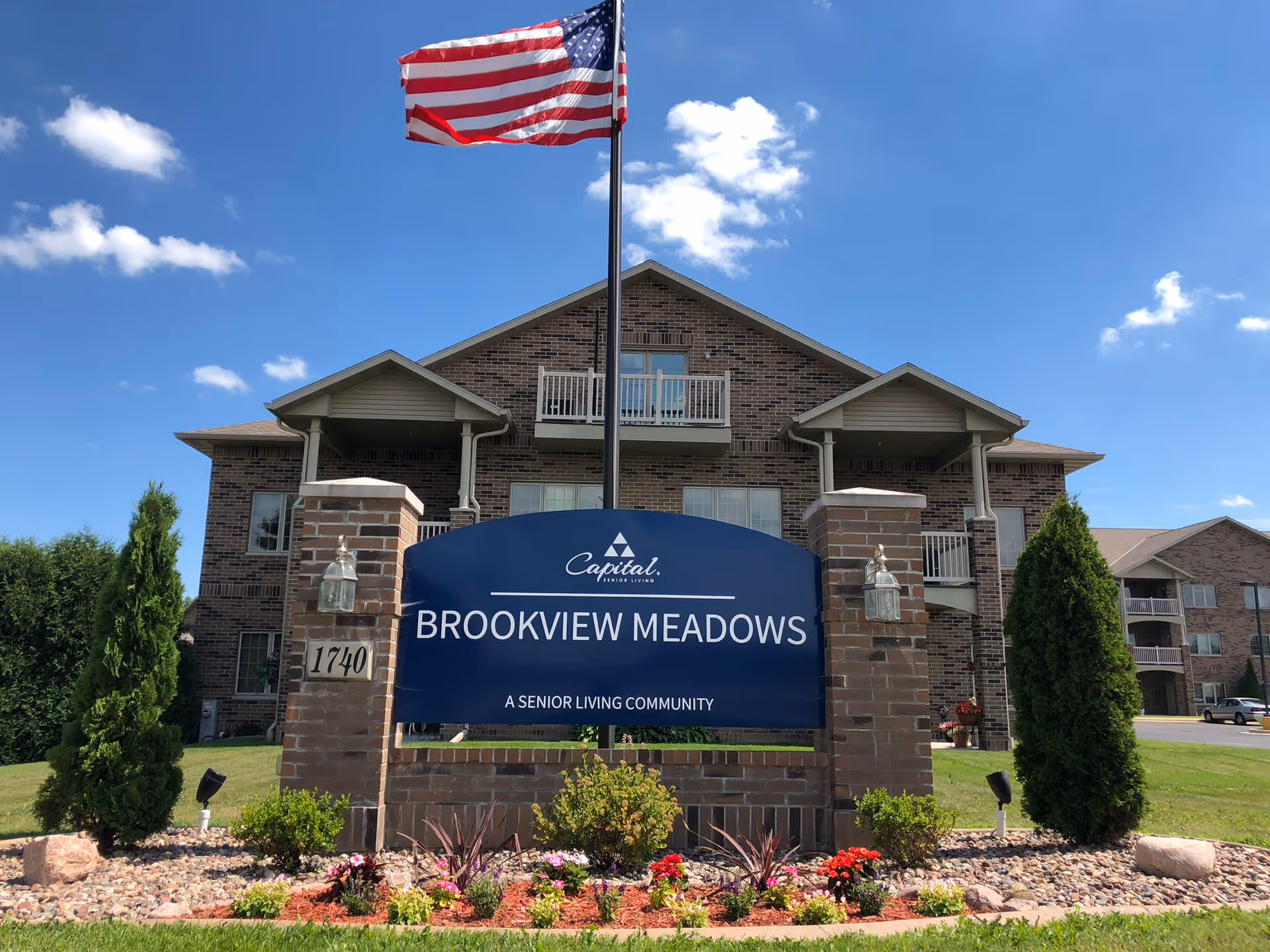 Exterior view of Brookview Meadows senior living community building with a large blue sign in front displaying the name and an American flag flying above. The building is made of brick with balconies and surrounded by green grass, trees, and a flower bed.