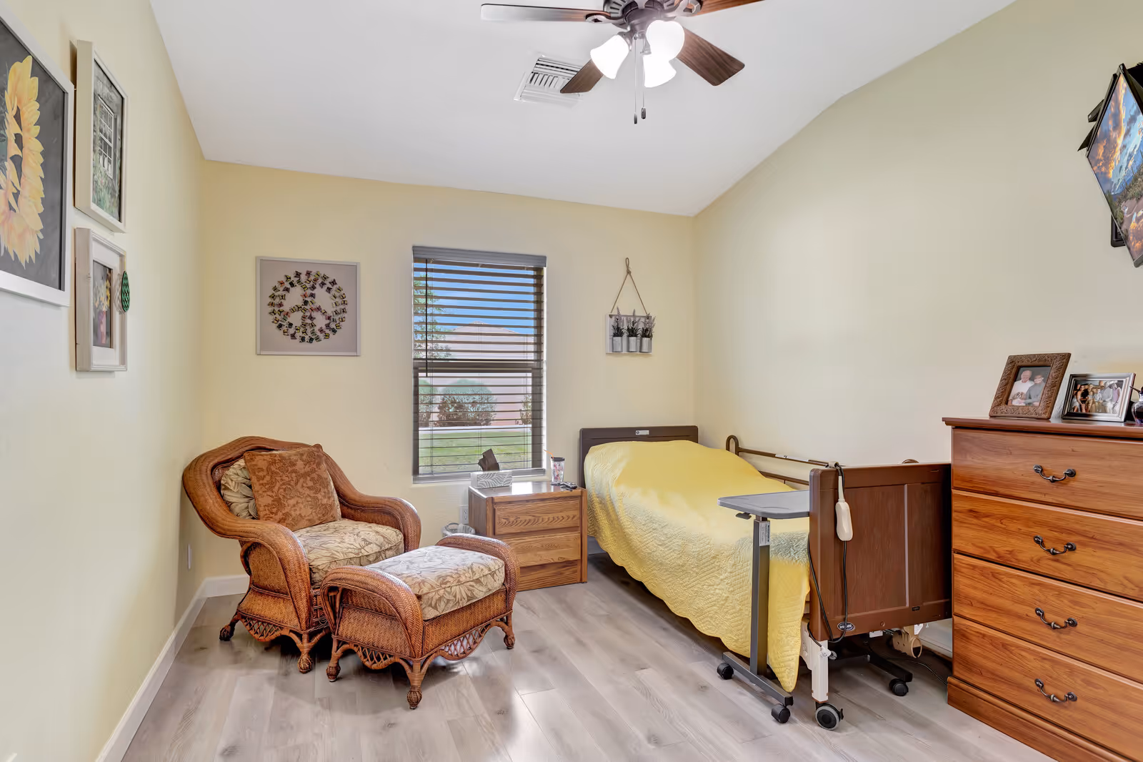 A cozy senior living bedroom with a single bed covered in a yellow quilt, a wooden dresser with framed photos, a wicker armchair with a matching ottoman, a wooden nightstand, and a window with blinds letting in natural light. The walls are painted light yellow and decorated with framed artwork and a small hanging plant. A ceiling fan with lights is mounted on the ceiling.