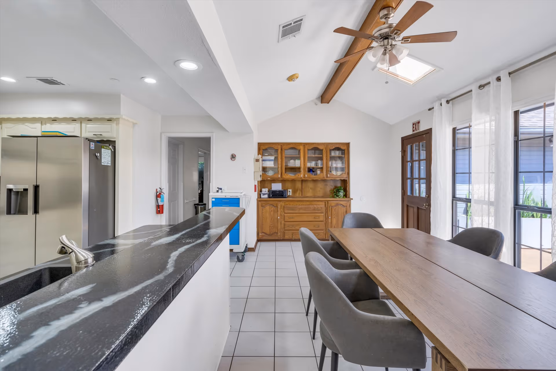 Interior view of a dining area with a wooden dining table surrounded by gray upholstered chairs. To the left is a kitchen counter with a black marble countertop and a stainless steel refrigerator. At the back, there is a wooden cabinet with glass doors and shelves. The room has white tiled flooring, a ceiling fan with wooden blades, and large windows with sheer white curtains letting in natural light.