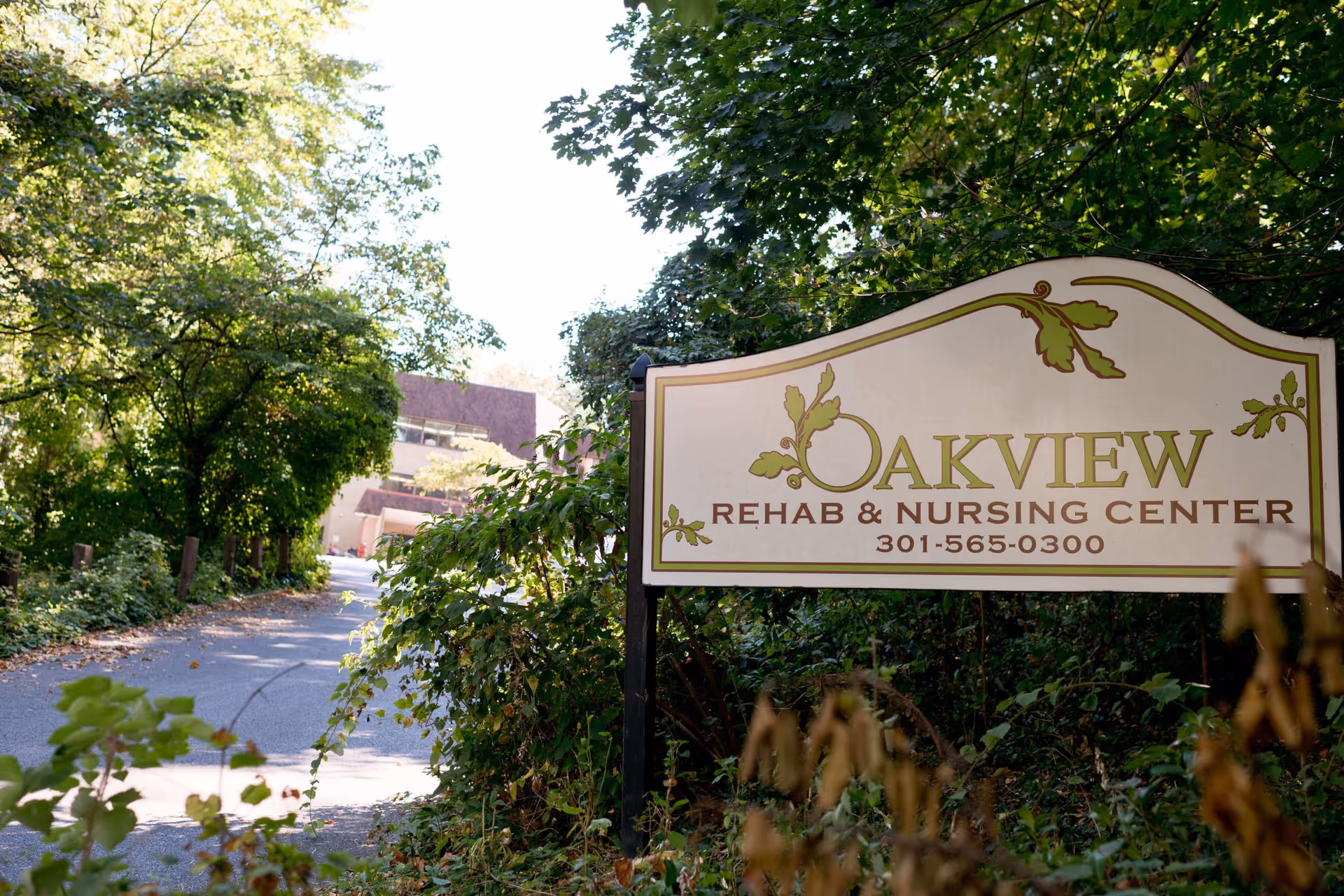 A sign for Oakview Rehab & Nursing Center with the phone number 301-565-0300, surrounded by green trees and bushes along a driveway leading to a building in the background.