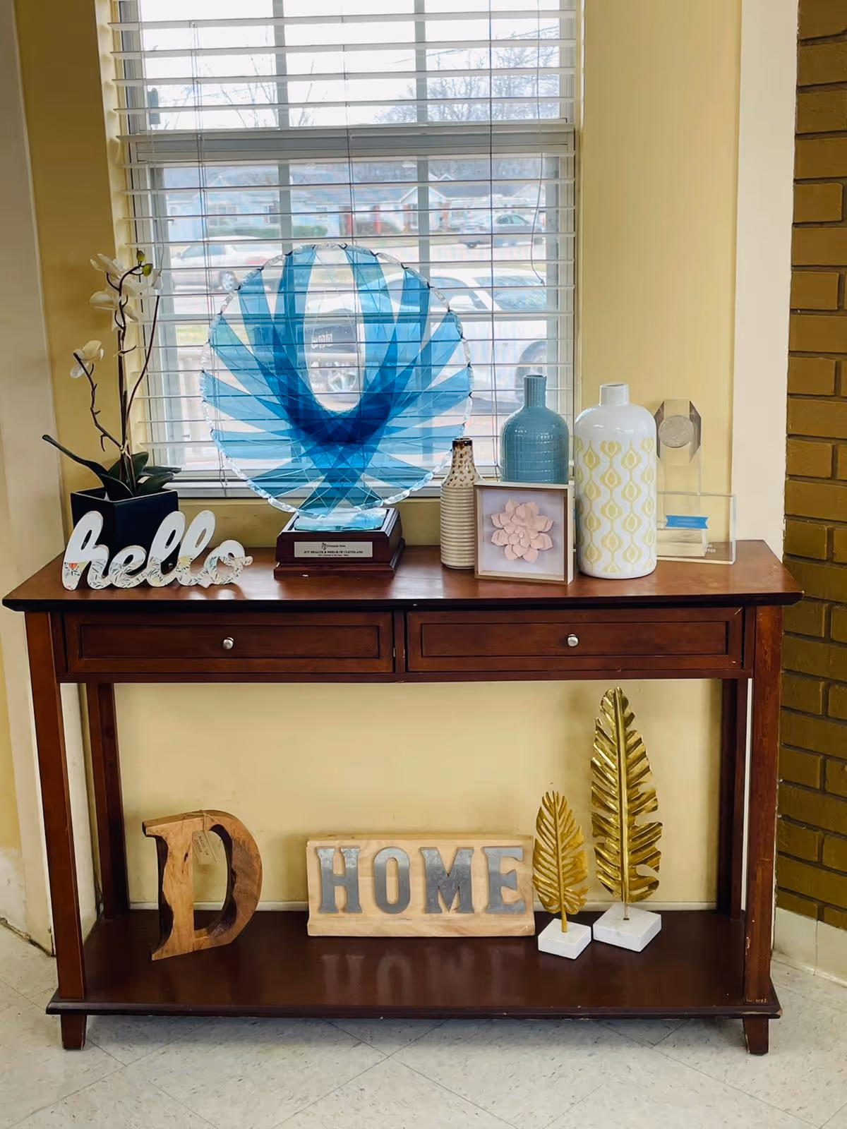 A wooden console table with two drawers placed against a wall under a window with white blinds. On top of the table are decorative items including a blue and clear glass award, a black pot with white flowers, a white decorative 'hello' sign, various vases, a framed pink flower artwork, and a clear acrylic award. Below the table are wooden and metal decorative pieces spelling 'HOME' and two gold-colored leaf sculptures.