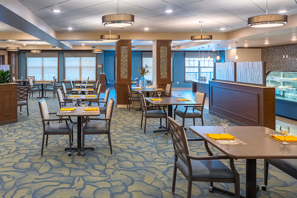 A spacious dining room in a senior living facility with multiple tables and chairs arranged neatly. Each table is set with yellow napkins and glassware. The room features blue walls, patterned carpet, modern light fixtures, and large windows allowing natural light to enter. A counter area with a display case is visible on the right side.