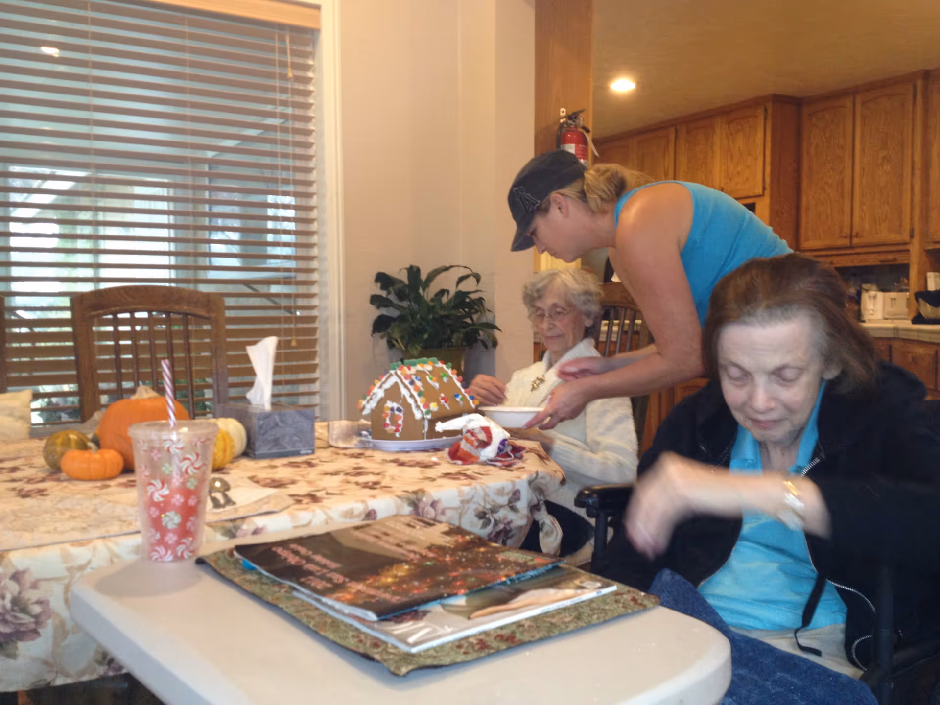 Three women gathered around a dining table in a homey kitchen/dining area with a gingerbread house, small pumpkins, and magazines on the table.