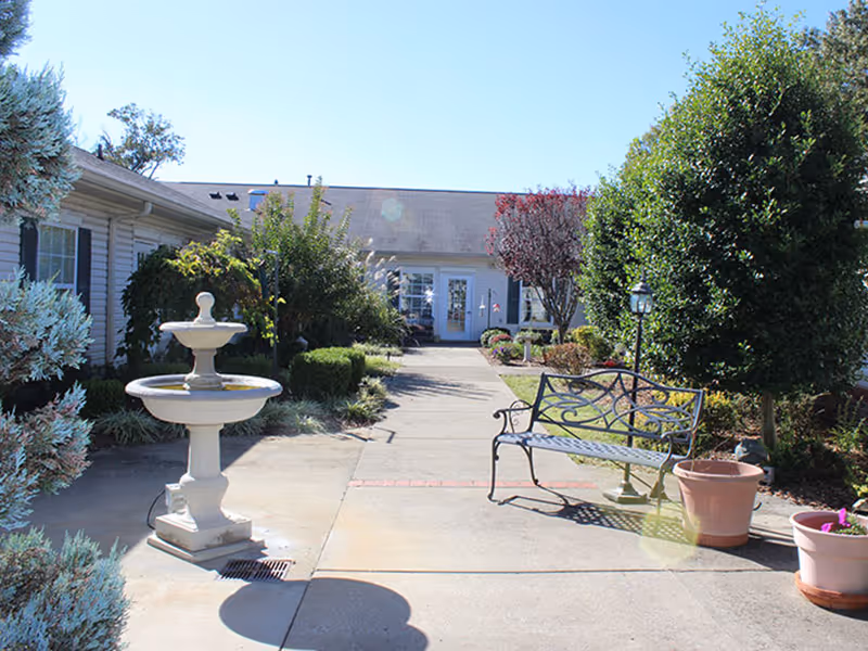 A well-maintained garden pathway leading to the entrance of Olive Branch Senior Living, featuring a decorative fountain, benches, and lush greenery.