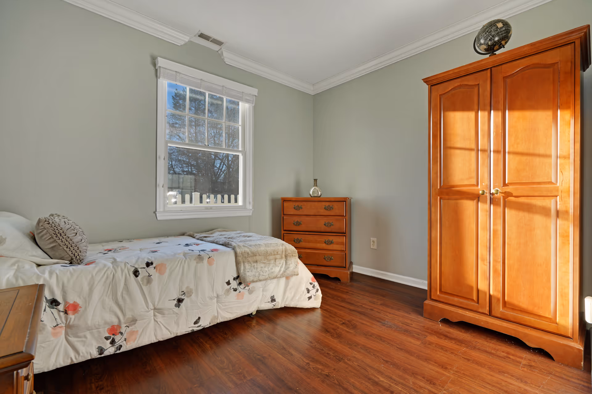 A simple bedroom with a single bed covered in a floral comforter and a round decorative pillow. The room has light green walls, a window with white trim letting in natural light, a wooden dresser with a decorative item on top, and a wooden wardrobe with a globe placed on it. The floor is a dark wood laminate.