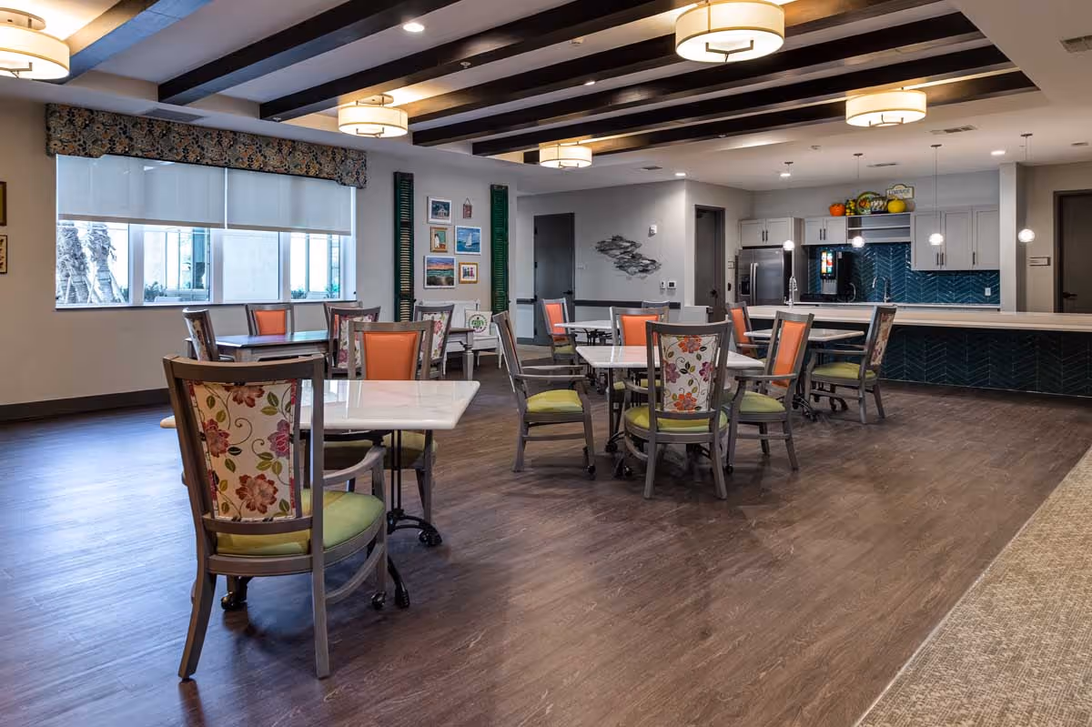 Open dining room with multiple tables, colorful upholstered chairs, and a kitchenette/serving counter in the background.
