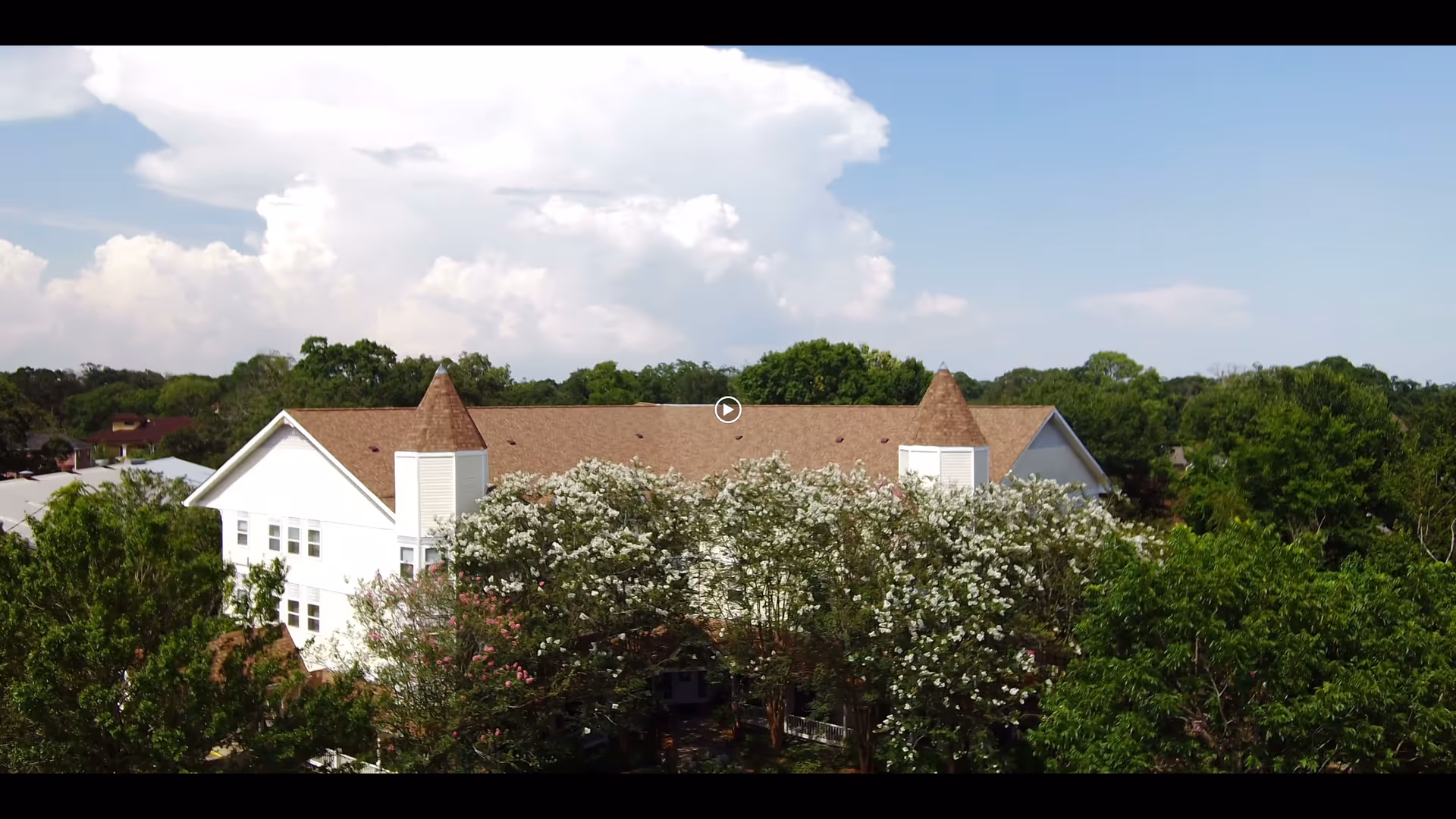 Exterior view of a large white building with a brown shingled roof and two turret-like structures, surrounded by green trees and flowering bushes under a partly cloudy sky.