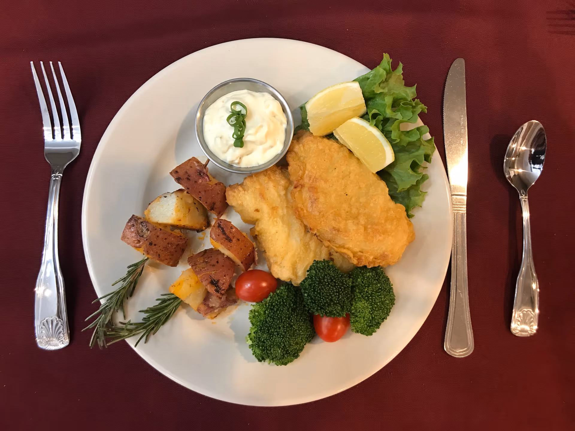 A white plate on a maroon tablecloth with a meal consisting of two pieces of fried fish, roasted potato chunks on rosemary sprigs, two lemon wedges on a bed of lettuce, two cherry tomatoes, broccoli florets, and a small metal cup of tartar sauce garnished with sliced green onions. Silver fork, knife, and spoon are placed on either side of the plate.