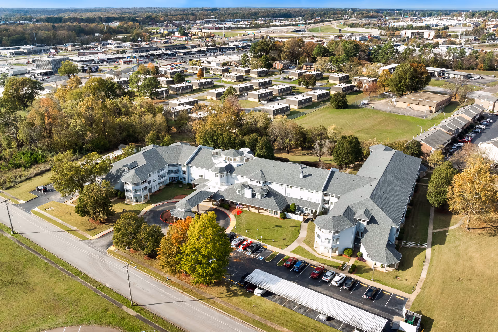 Aerial view of Jackson Meadow - A Provincial Senior Living Community, showing a large multi-wing building surrounded by trees and parking lots, with a suburban neighborhood and roads in the background under a clear sky.