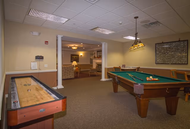 Interior view of a senior living facility game room featuring a shuffleboard table on the left and a pool table on the right. The room has beige walls, carpeted floor, and a ceiling with recessed lighting. In the background, there is a seating area with armchairs, a lamp, and a fireplace. A framed picture hangs on the wall above the pool table.