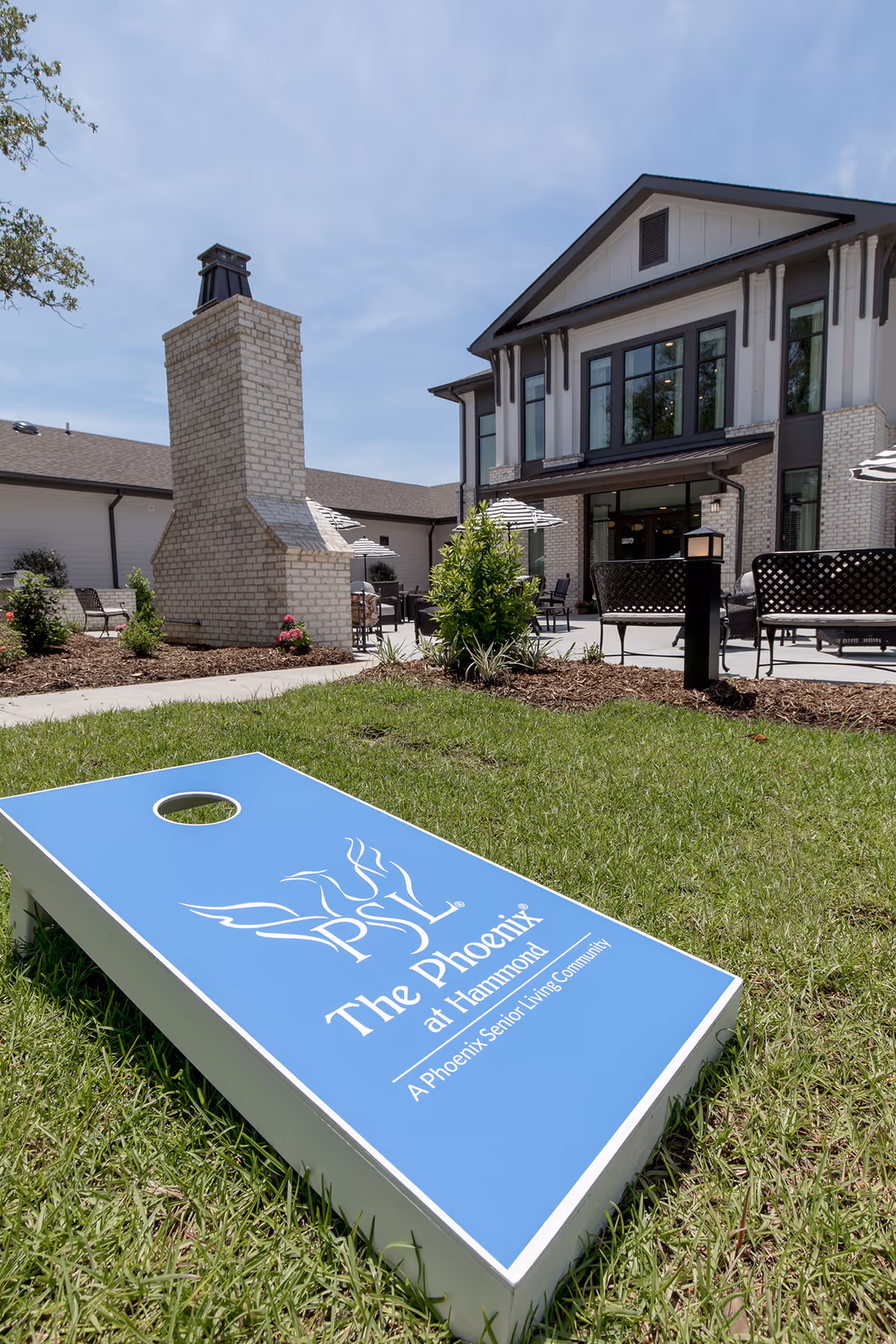 Outdoor patio area of The Phoenix at Hammond senior living community featuring a brick outdoor fireplace, patio tables with umbrellas, chairs, and a cornhole game board on the grass in the foreground.