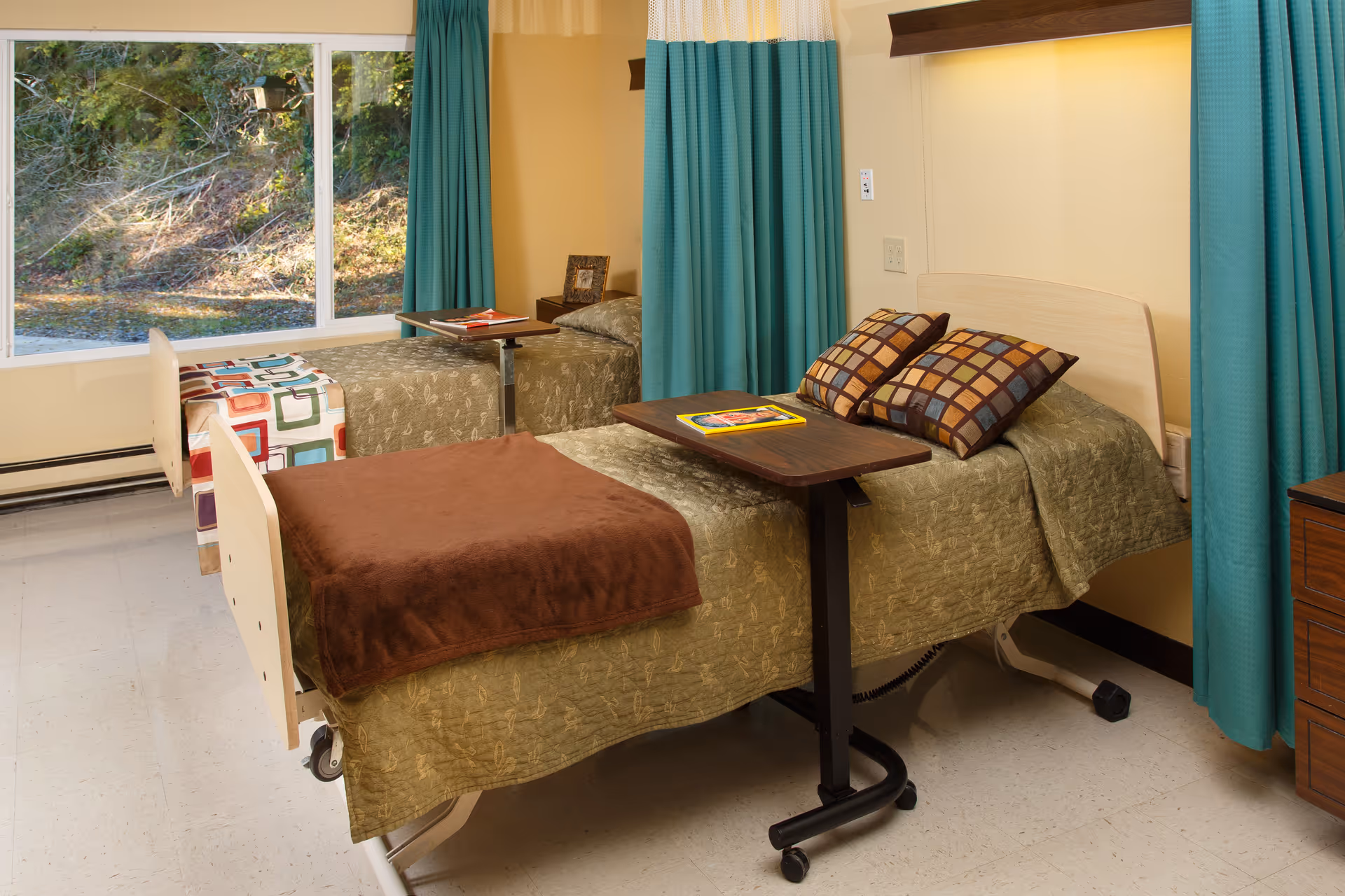 A room with two hospital-style beds separated by a teal privacy curtain. Each bed has a green bedspread and a brown blanket, with pillows on the bed closest to the camera. There are overbed tables on each bed, one with a book and the other with a red item. A large window shows an outdoor view with trees and bushes.